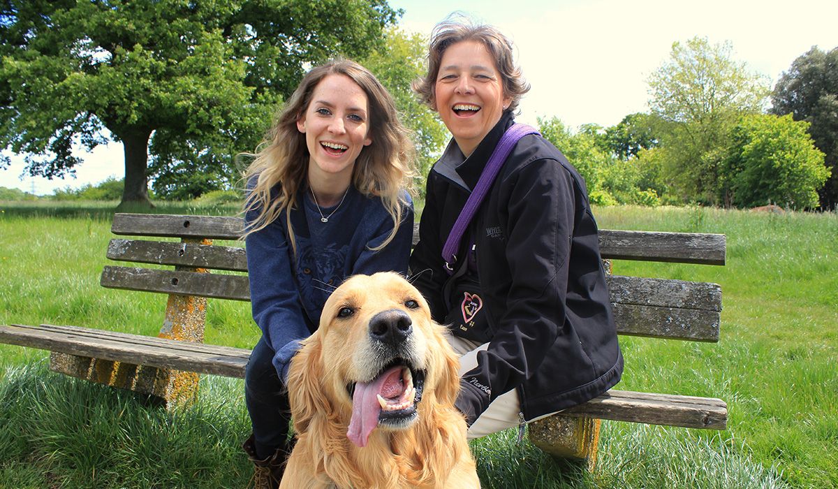 Two women with large smiles are sitting on a bench in the countryside. In front of them sits Bailey, a happy Golden Retriever  with his tongue hanging out