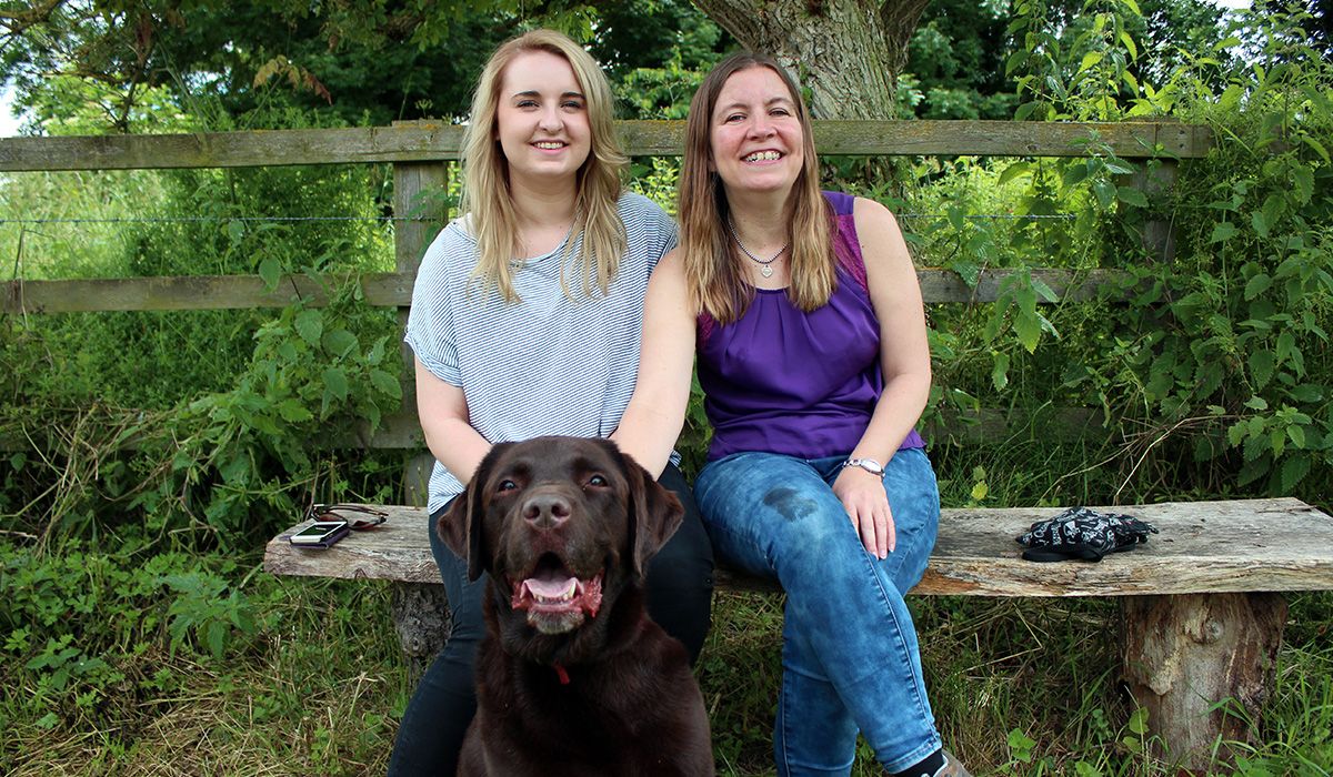 Two women are sitting on a bench. In front of them is Bertie, the Chocolate labrador - a large, dark brown dog.