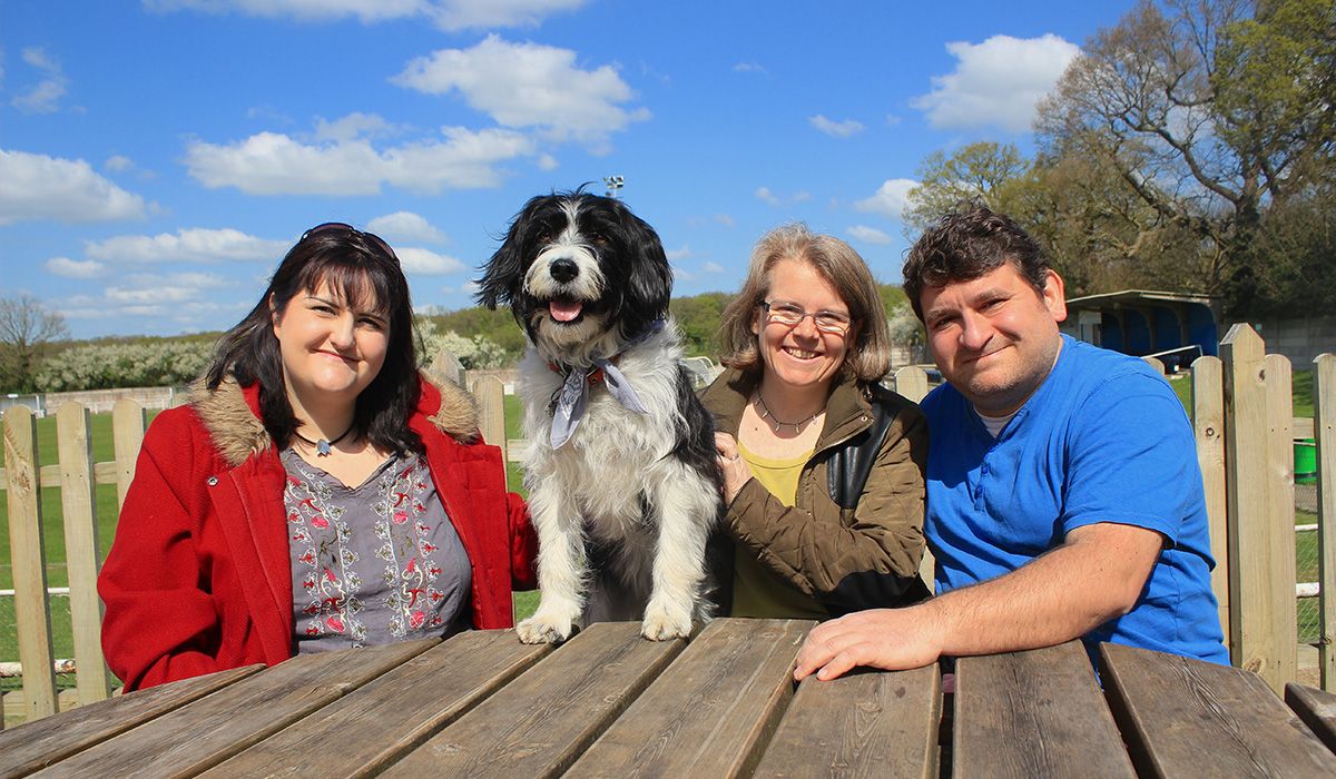 Three people and a shaggy, black and white dog are sitting at a picnic table on a sunny day