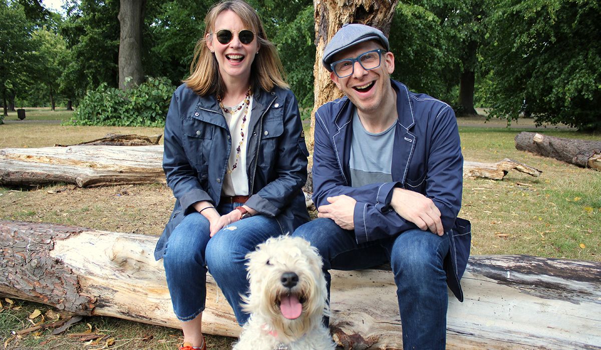 Two smiling people are sitting on a log in woodland. They have broad grins and in front of them is a white, curly-haired dog with their tongue out