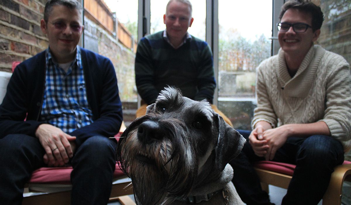 A grey dog with an impressive moustache is in the foreground. Behind him sit 3 men. 
