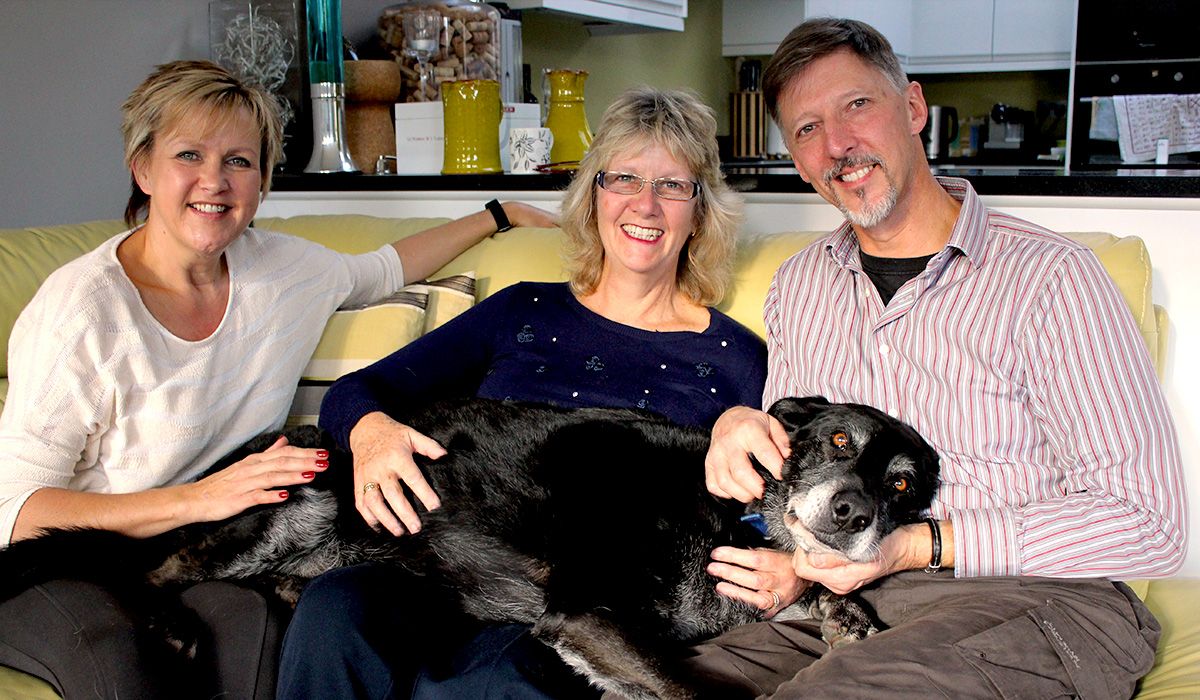 three people on a light yellow couch with a dog lying on top of them all