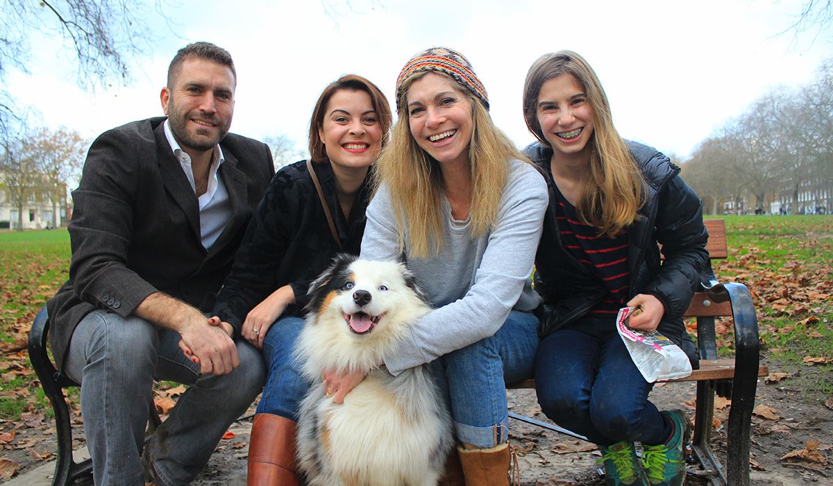 Four happy people are squeezed onto a park bench. In front of them is a very fluffy, mostly white dog with a big smile.