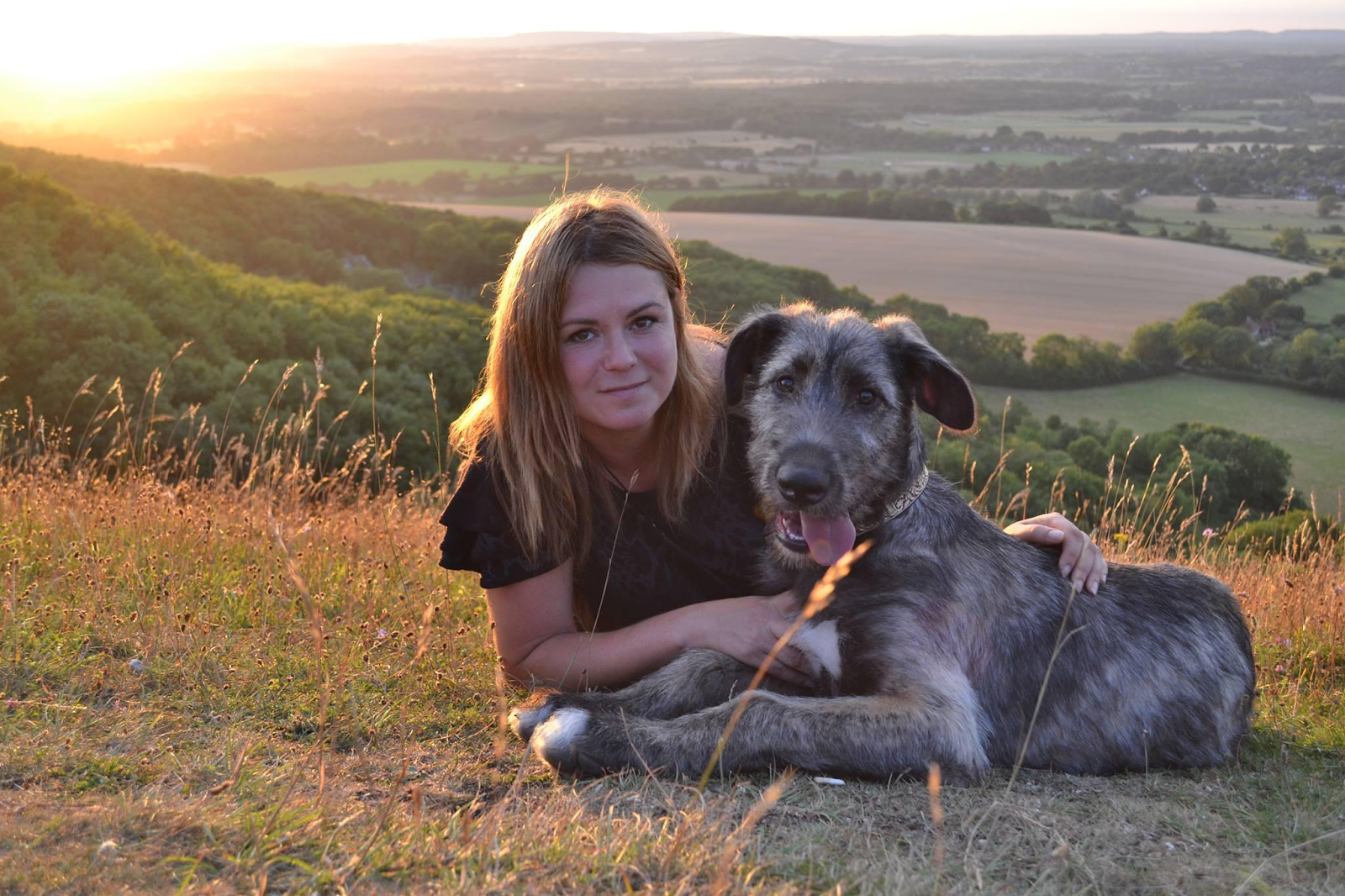A woman and a dog who is nearly her size are lying together on a hill at sunset.
