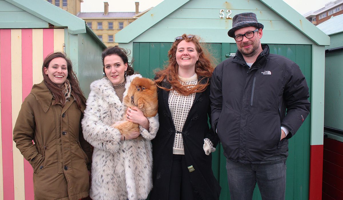Four people and a small, ginger dog are standing by the seaside on a windy day
