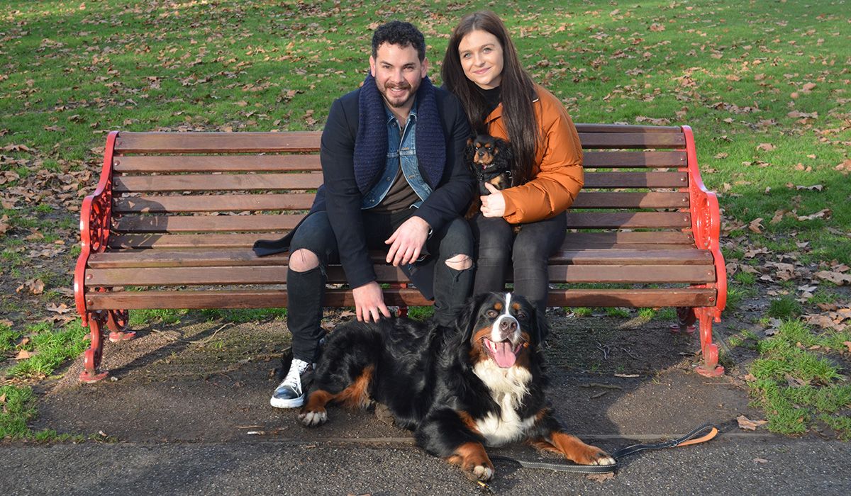 Two people are sitting on a bench in a park. A large black, white and tan dog lies at their feet, a small black and tan dog is on their lap.