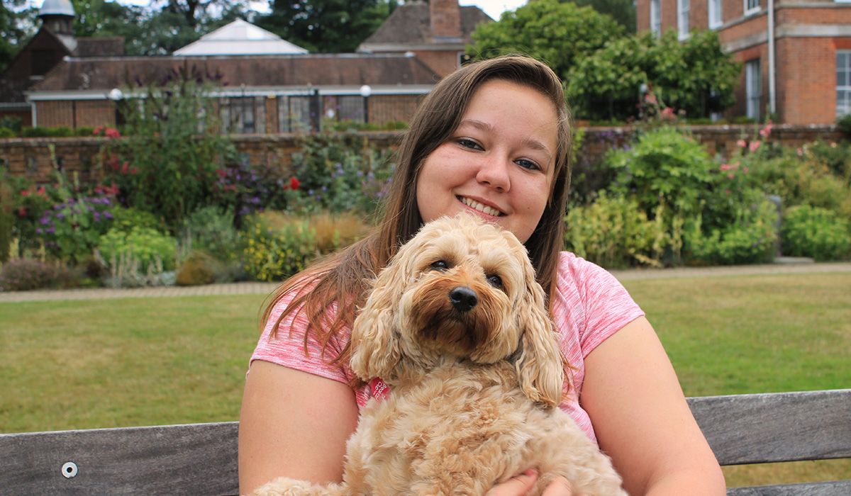 Victoria is sitting on a bench in formal gardens. On her lap is a blonde, curly haired dog who she is cuddling lovingly.