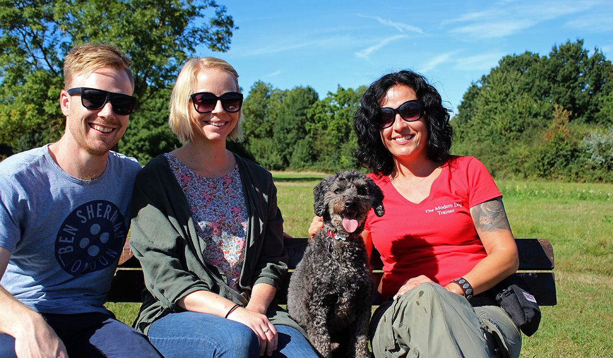 Three people and a black, curly-haired dog are sitting on a bench in a park on a beautiful, sunny day