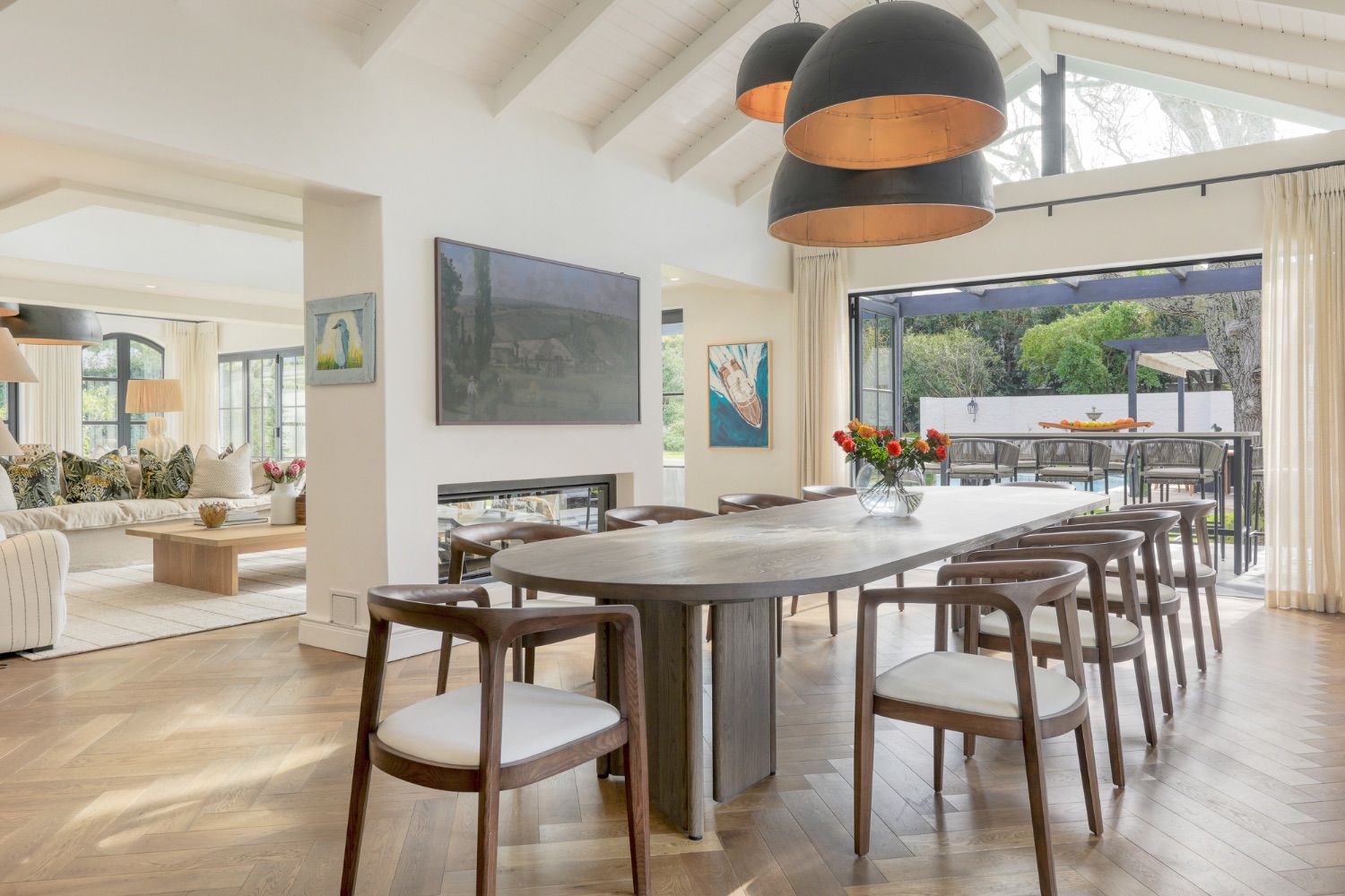 An open-plan dining and living room with an oval oak table, black pendant lights and doors opening onto a garden terrace