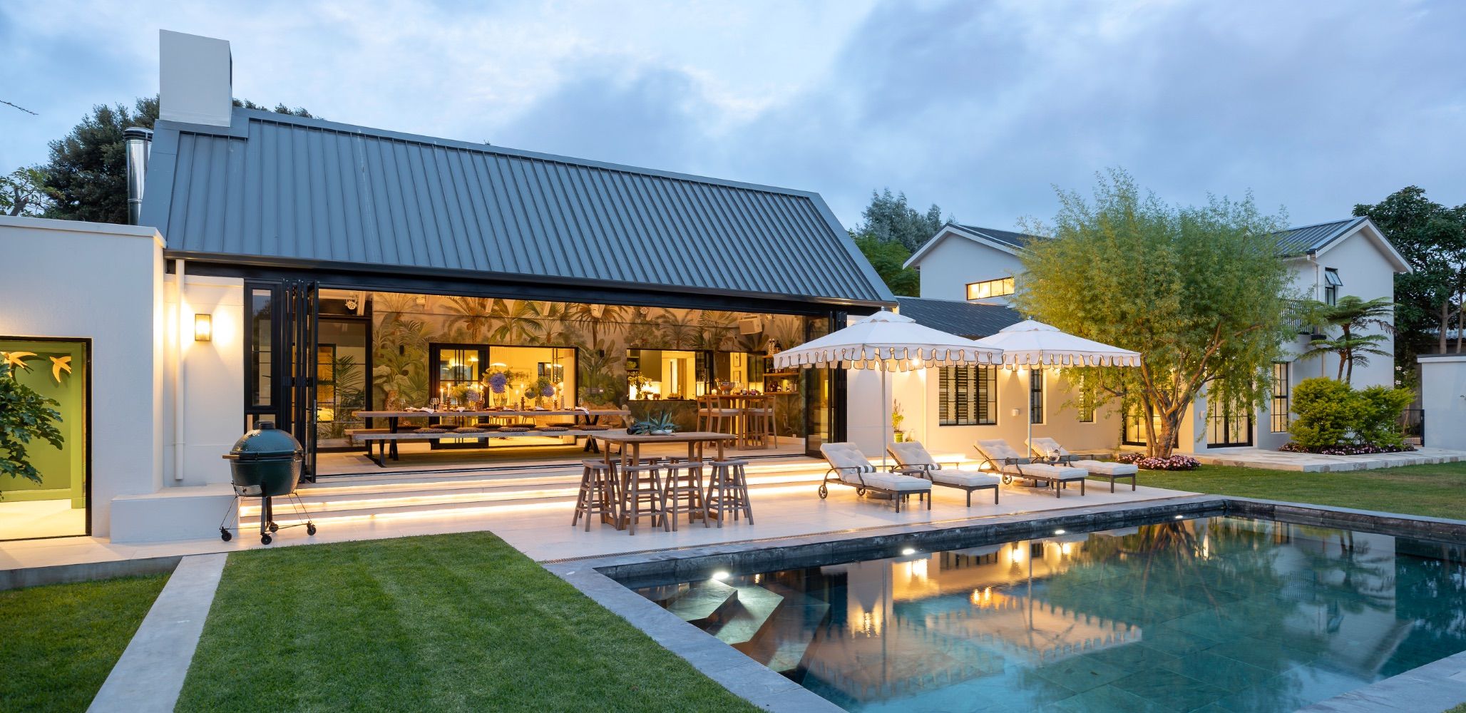 A white villa with a dark pitched roof, a covered terrace and a pool at dusk
