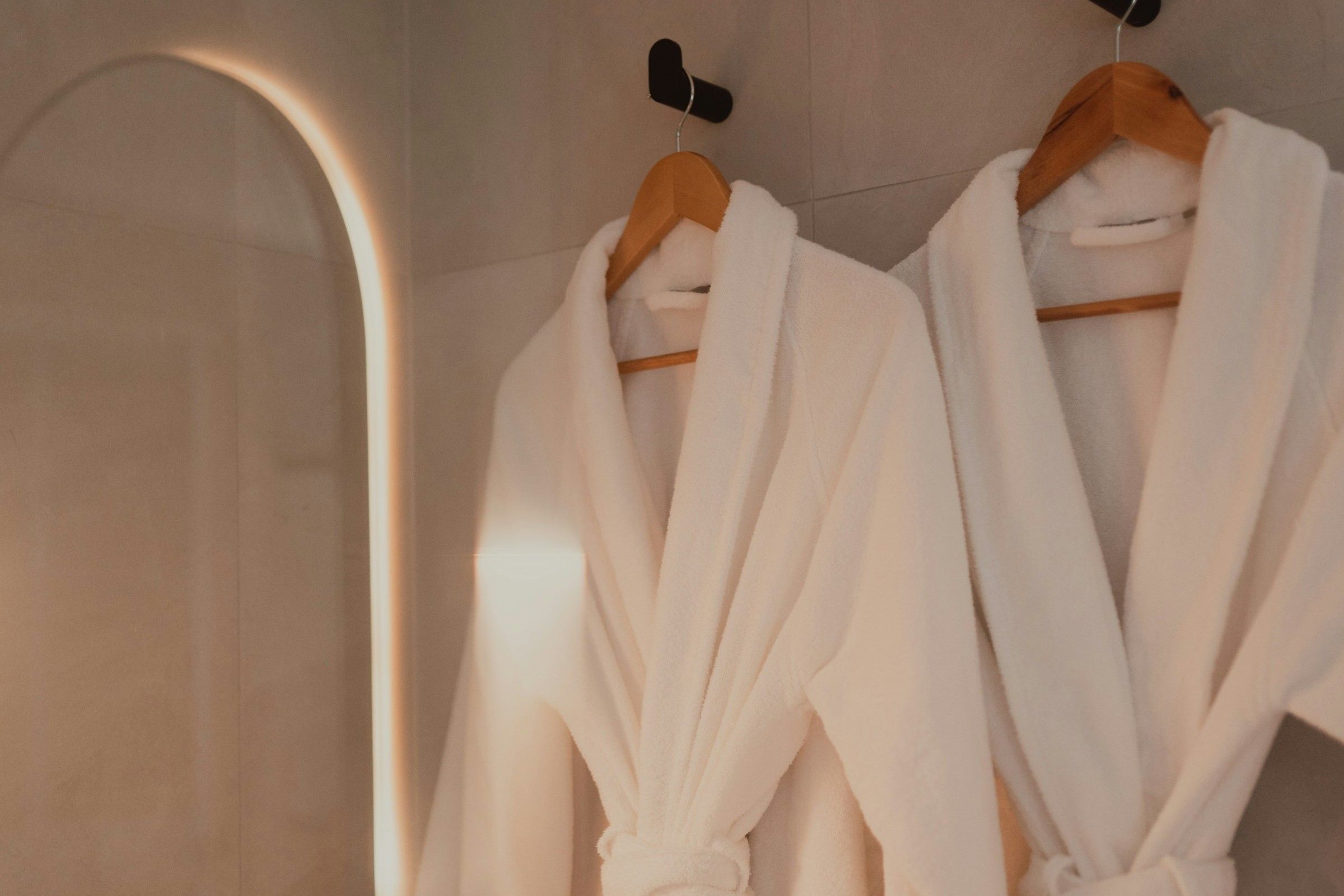 Two white hotel bathrobes hanging on wooden hangers in a bathroom