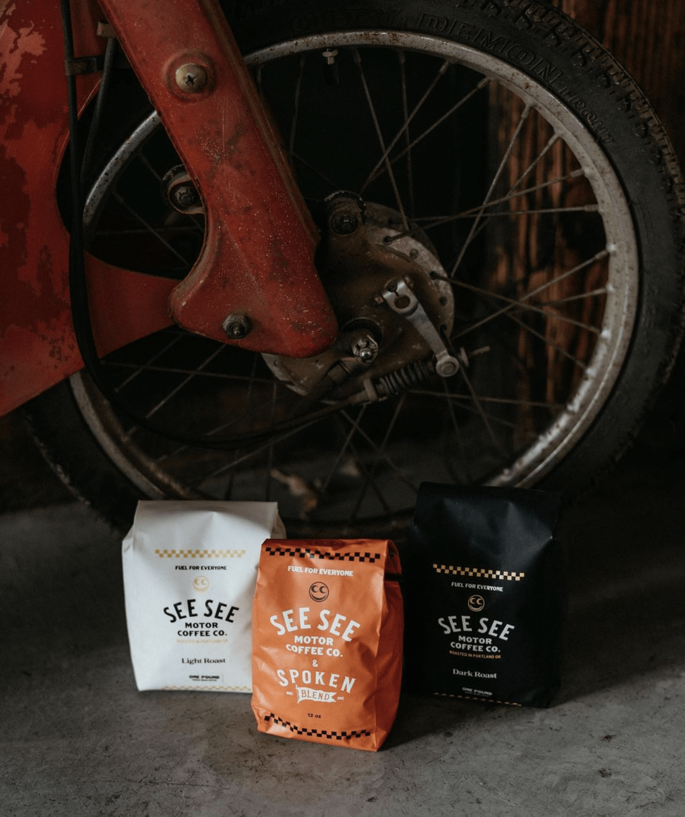 Three See See Motor Coffee bags of coffee beans sitting on a concrete floor next to a motorcycle wheel.
