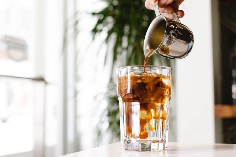 Espresso being poured into a glass of ice water, in a room filled with light and plants