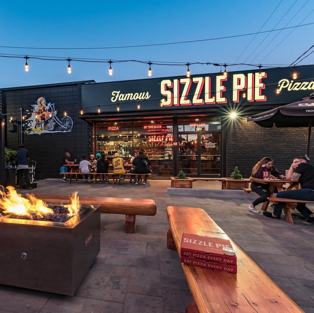 The front of a Sizzle Pie restaurant with outdoor seating, and benches around a fire pit, with red pizza boxes in the foreground