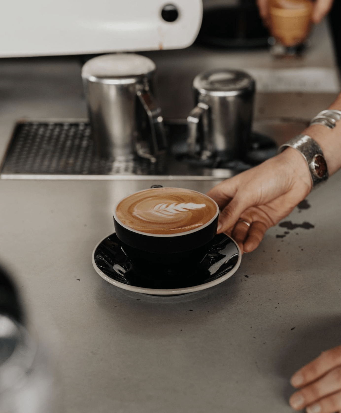 A close up of a barista hand serving a beautifully poured latte in a black cup on a saucer. 