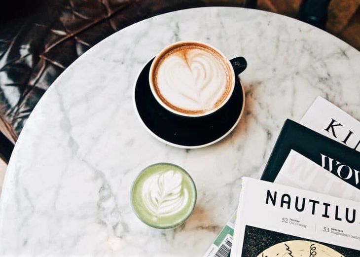 A top-down view of a latte with latte-art in a black cup, on a black saucer, next to a matcha latte and a pile of magazines, all on a marble table top