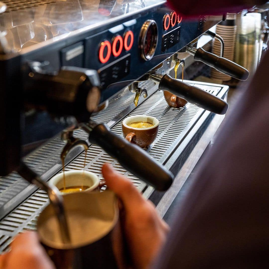 A close-up of an espresso machine with 3 groups, while a barista steams milk in the foreground