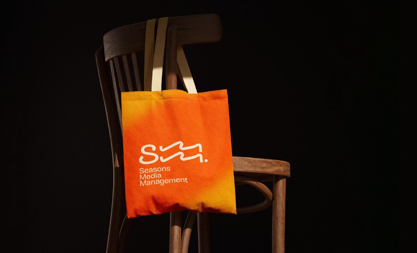 Orange Seasons Media tote bag resting on a wooden chair against a warm beige wall. Brand Design by Efem Studios