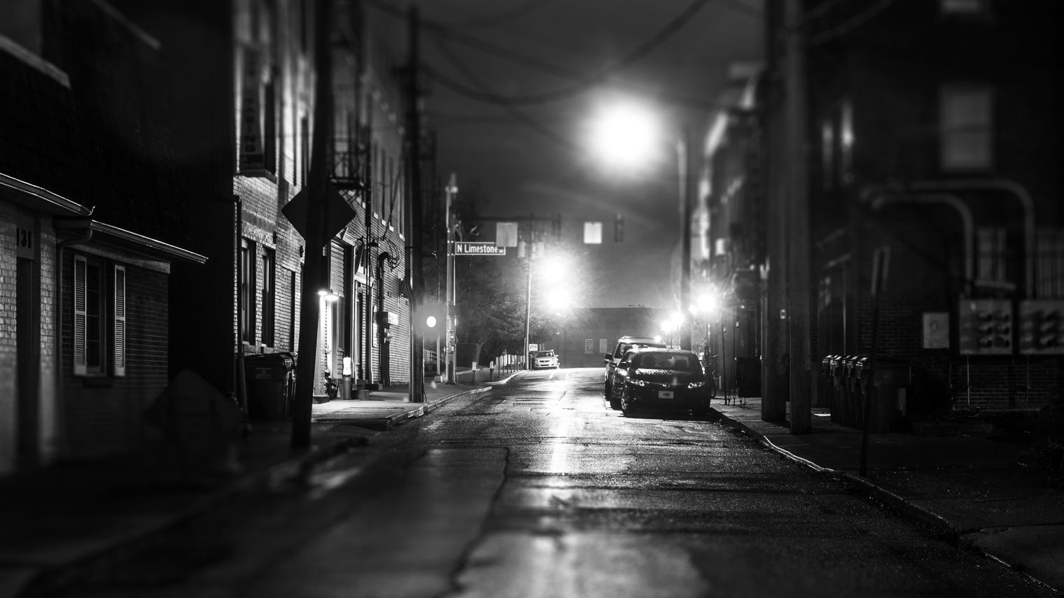 A suburban, nighttime vista: a dark stretch of road in town, lined by brick buildings and lit intermittently by street lamps, wet from the rain.