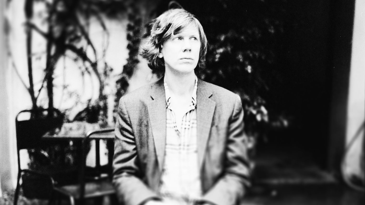 Thurston Moore sitting down, wearing a suit jacket and a button-down shirt, looking to the right. 