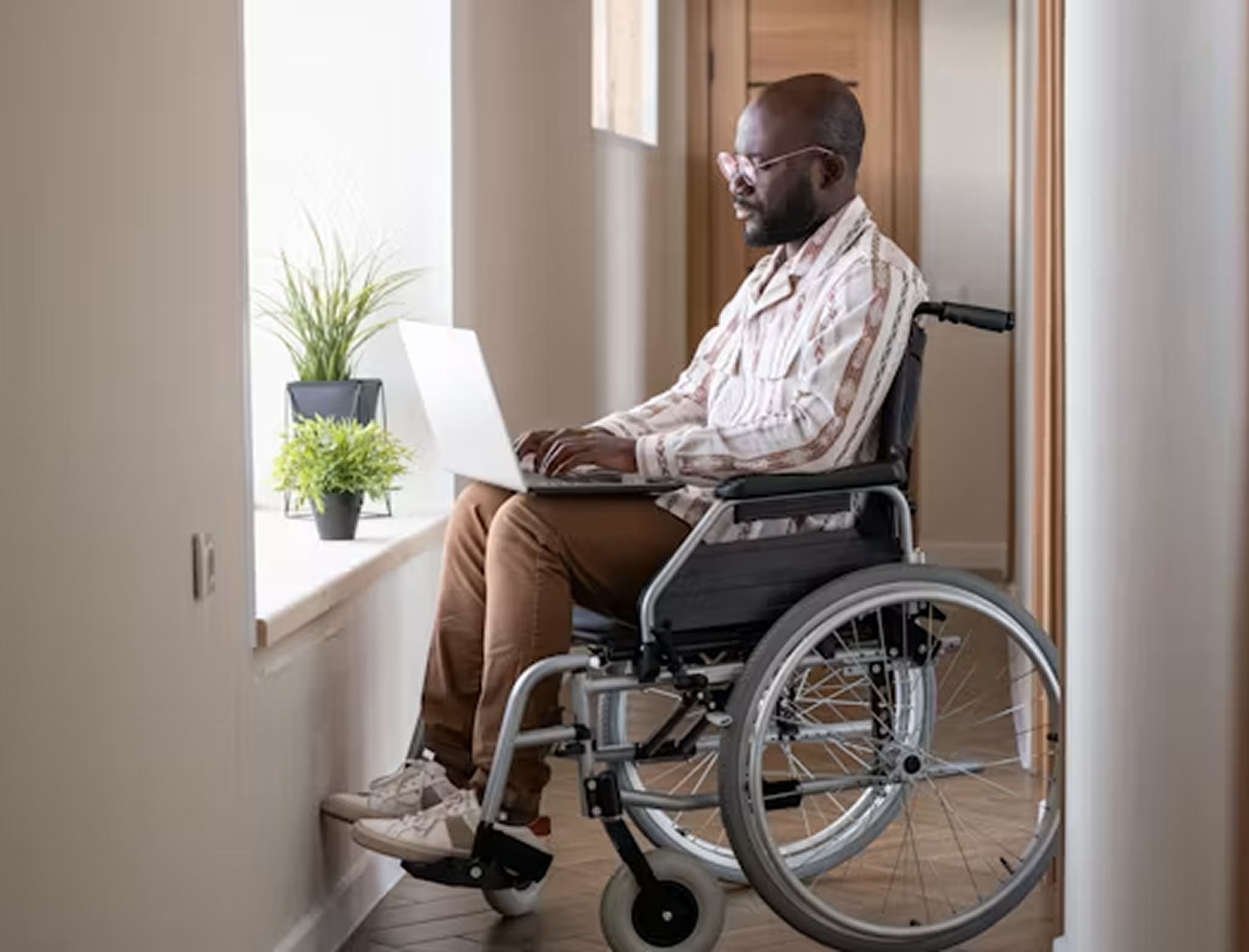 Man using wheelchair working on laptop near bright window