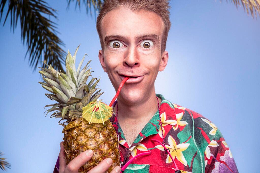 Mand i blomstret hawaii-skjorte som drikker af en ananas gennem et sugerør. Han har store øjne.