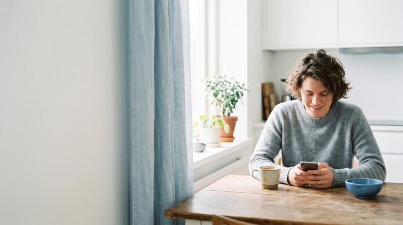 A man smiling at his phone over coffee at a sunlit kitchen table