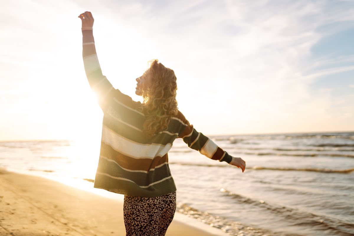 Vrouw staat met open armen op het strand in tegenlicht