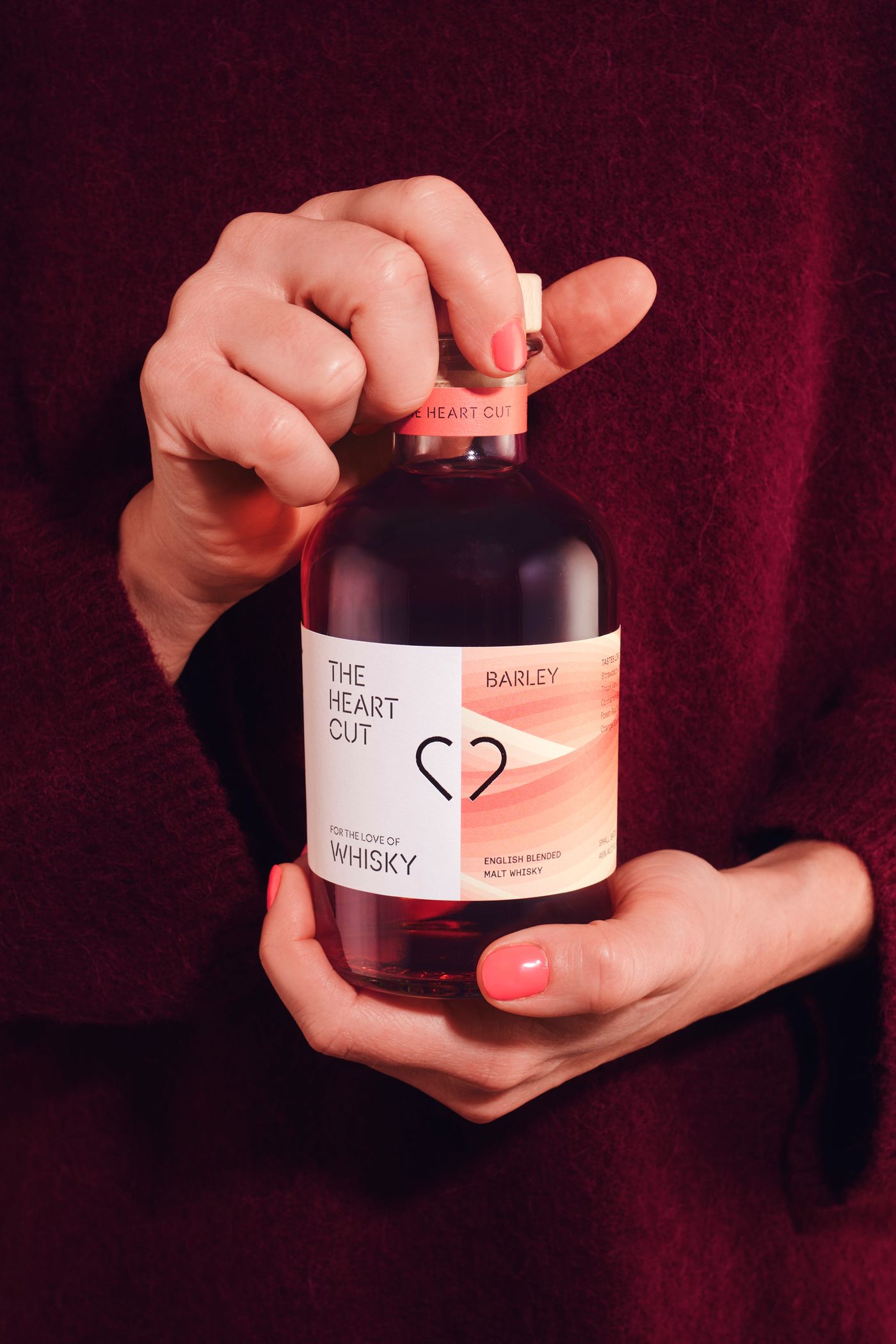 In front of a burgundy jumper, a woman's hands hold a bottle of red-tinged The Heart Cut whisky giving a Christmas vibe