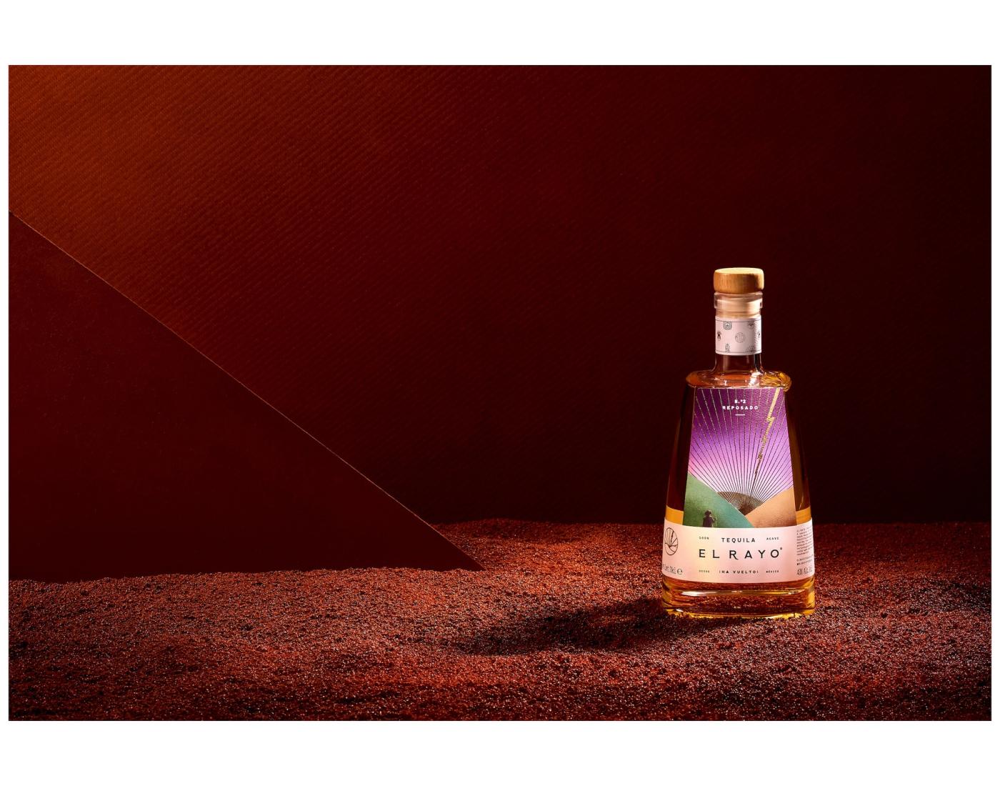Bottle of El Rayo Reposado tequila standing on red sand against a dramatic dark red backdrop