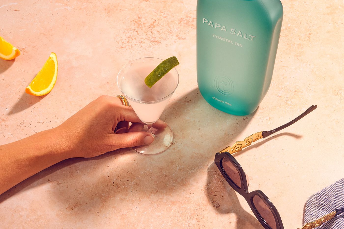 A beachy surface. On it a woman's hand pushes a v-shaped cocktail glass, around her sunglasses, two slices of orange, and a bottle of Papa Salt Gin, while the afternoon sun casts long shadows.