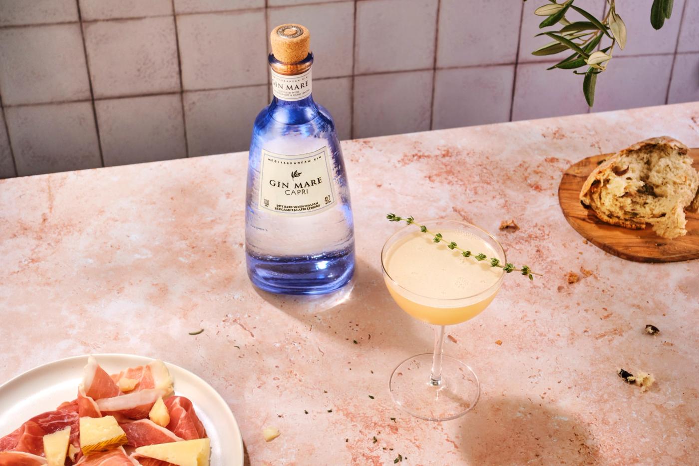 A bottle of Gin Mare, with two plates of tapas, and a thyme-garnished cocktail. The surface is pink marble, the backdrop is faded tile, a sprig of Mediterranean lean hangs in the corner.