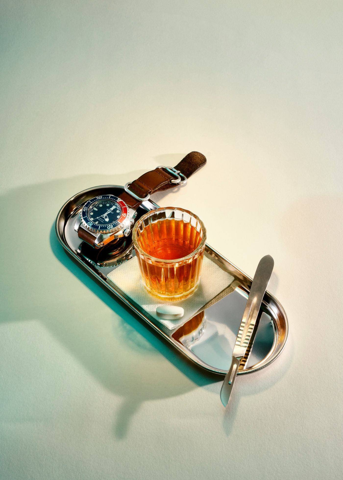 A shot of whisky sits on a surgeon's tray. Next to it is a Seiko divers watch,. a scalpel and a tablet.