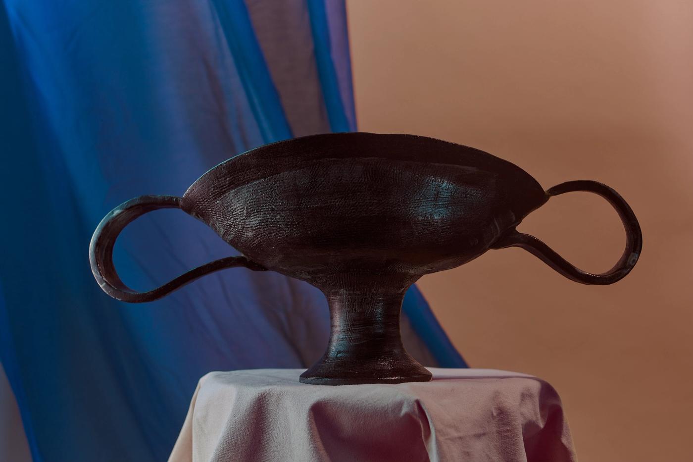 A Greco-Roman style urn sits on a plinth with a flowing blue curtain behind it.