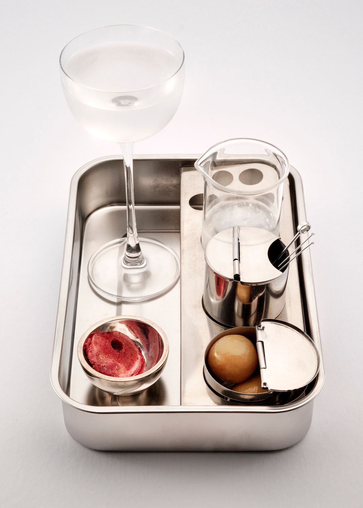 Cocktail glass, glass beaker and hinged surgical dishes holding olives and a real trimmed piece of the artist's femur, arranged on a surgical tray, styled as "Garnish"