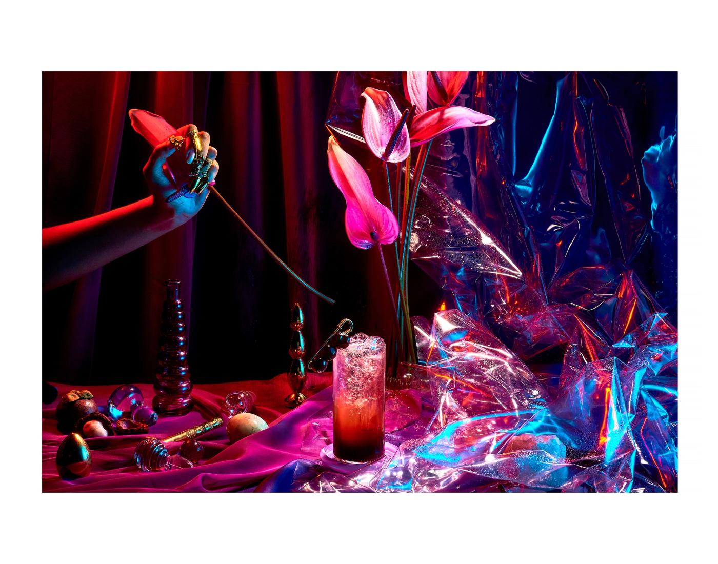 A cinematic, surreal set with coloured gel lighting shows a hand pulling a Cala Lilly, a cocktail stands in the middle, a blue-tinged plastic sheet is crumpled to the right, while an assortment of eggs are on the left