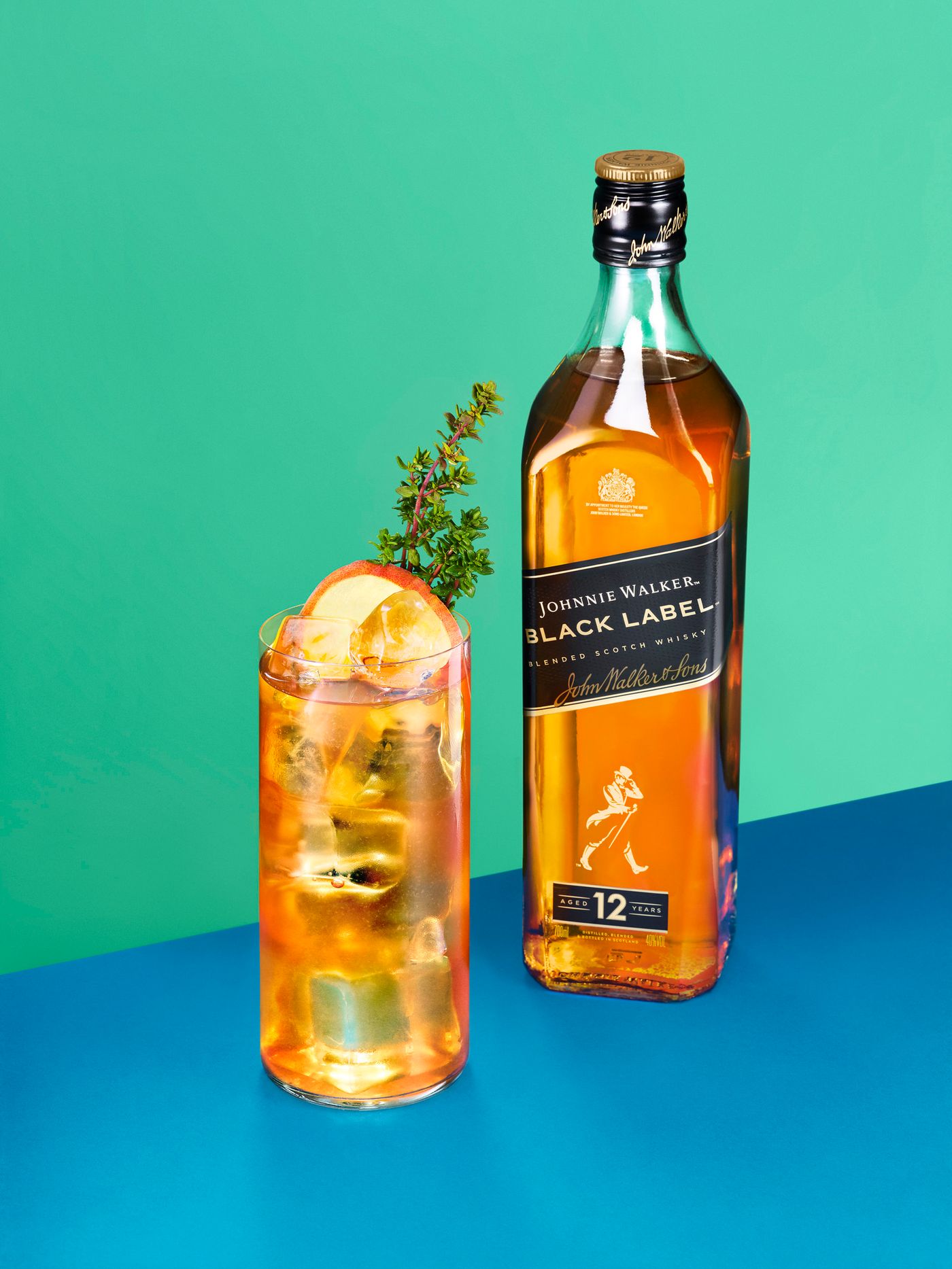 An apple-garnished whisky cocktail stands next to a Johnnie Walker Black Label  bottle on a colour-blocked green and blue set
