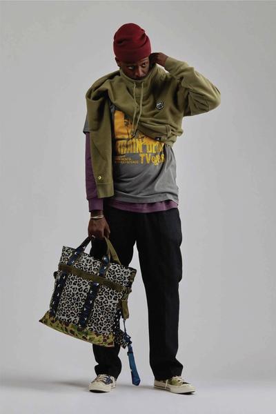 Man wearing chuck 70s, black chinos, green sweatshirt w gorilla embroidery around his waist, black bomber / hoody and a maroon watch cap beanie. Brain Dead x Converse collaboration.