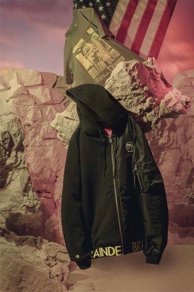 An image of a black bomber jacket perched in the desert. A tshirt and an american flag stand in the background.  Brain Dead x Converse collaboration.