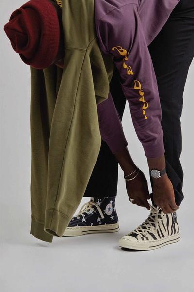 Man tying his shoes wearing chuck 70s, black chinos, green sweatshirt w gorilla embroidery around his waist, Maroon long sleeve tee and a maroon watch cap beanie. Brain Dead x Converse collaboration.