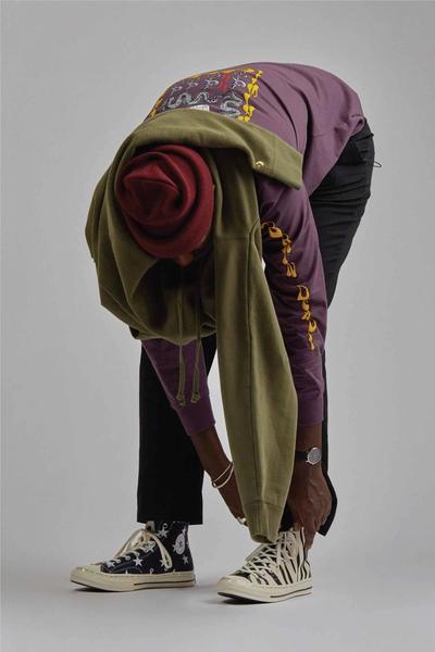 Man wearing chuck 70s, black chinos, green sweatshirt w gorilla embroidery around his waist, black bomber / hoody and a maroon watch cap beanie. Brain Dead x Converse collaboration.