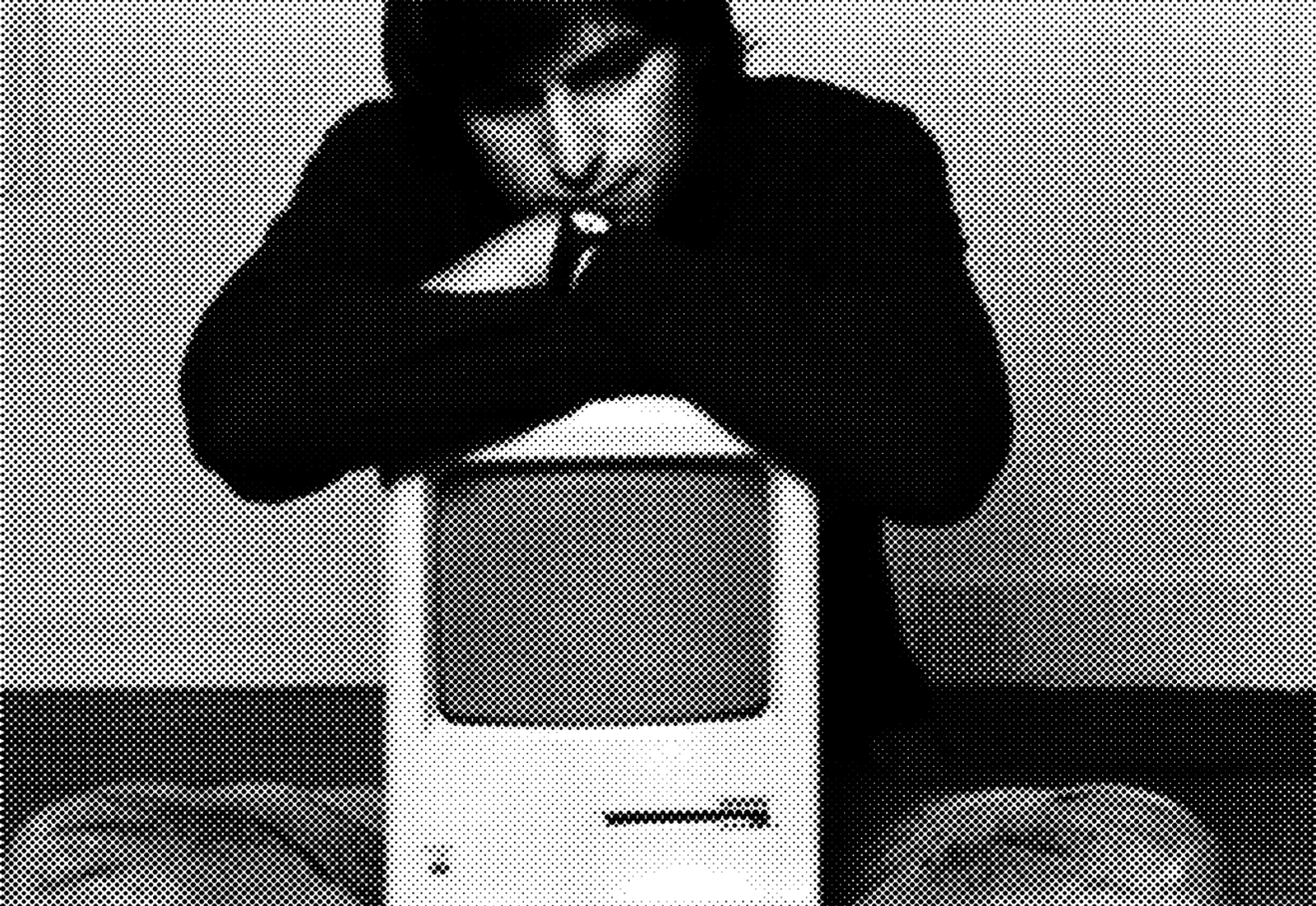 Black and white halftone portrait made of dots. A man with dark, tousled hair looks downward in thought, his chin resting on his folded arms, which lean on top of a small vintage compact computer. He wears a dark sweater and a wristwatch. The dots grow dense and bold in the shadows of the sweater and hair, and thin out to a light texture across the face, the computer's casing, and the background wall.