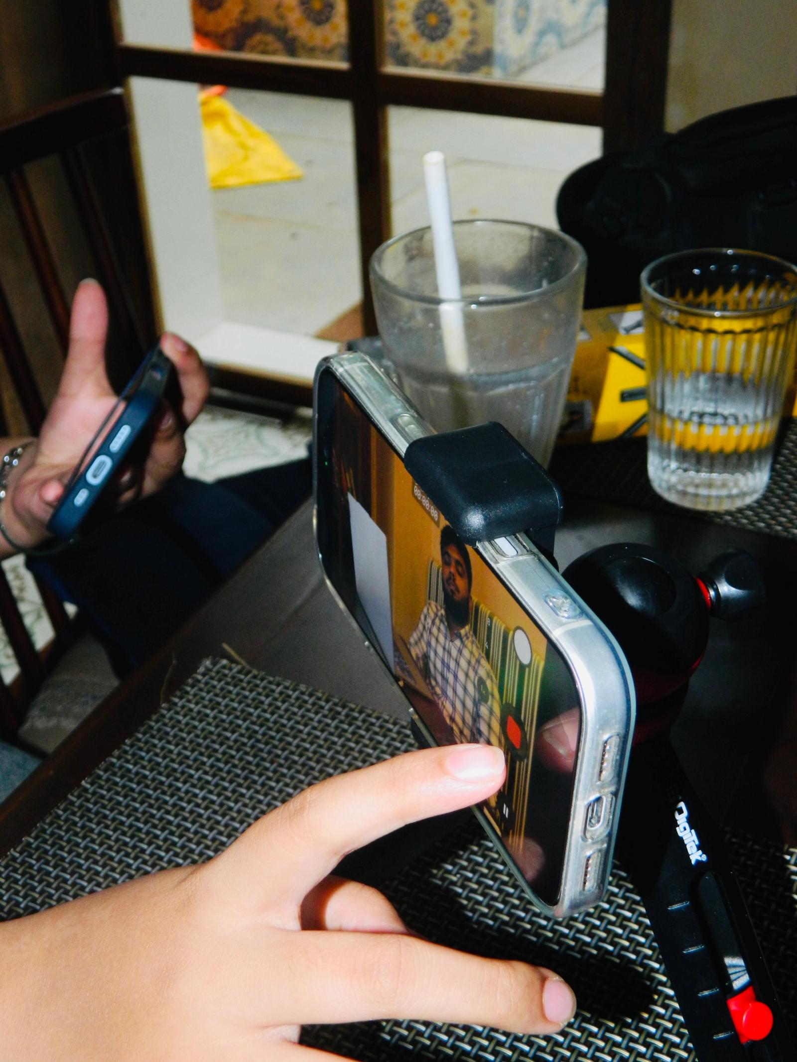 A hand points toward a smartphone mounted horizontally on a small black tripod on a table. The phone's screen shows a video recording in progress of a man in a plaid shirt. In the background, there are two drinking glasses and another person holding a phone.