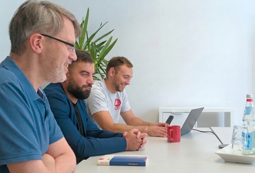 Drei Mitarbeiter von VP-Systeme sitzen an einem Tisch. Sie scheinen amüsiert über etwas zu sein. Im Hintergrund läuft ein Video-call auf einem Fernseher an der Wand.