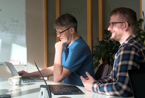 Zwei Softwareentwickler sitzen amüsiert nebeneinander mit ihren Laptops an einem Tisch in einem Meetingraum. Im Hintergrund ist ein Whiteboard zu sehen, auf dem mit Visualisierungen zu Maschinenlernen-Algorithmen gemalt wurden sind. 