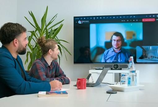 Depicted is a job interview situation, with two employees sitting beside each other in a meeting room, looking at a big television on the wall that displays another participant in a video call.