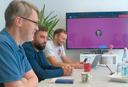 Three VP-Systeme employees sitting at a table, with a video call running on a television on the wall. They seem to be amused about something.