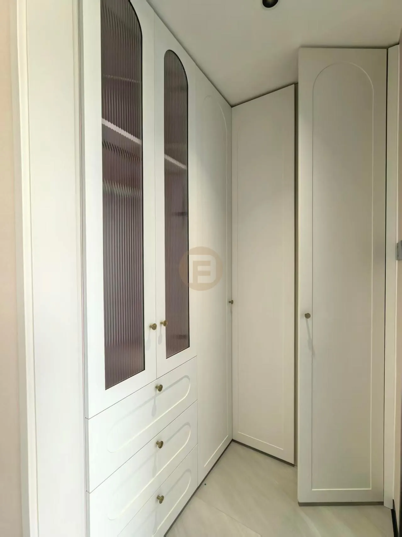 Cream arched wardrobes with reeded-glass doors and brass knobs lining a walk-in closet corner