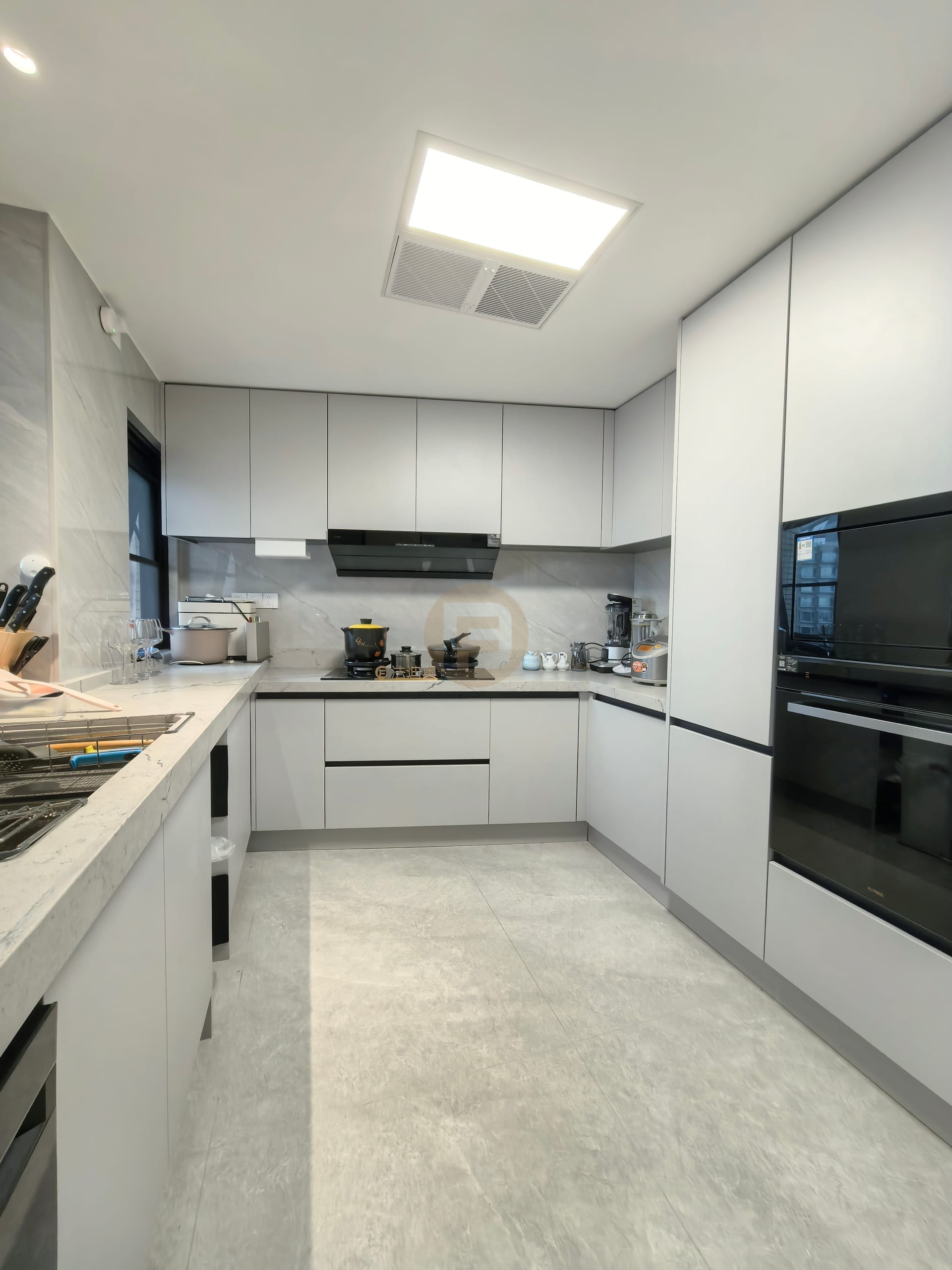 U-shaped kitchen in pale grey stainless steel cabinetry with black built-in ovens and marble-look splashback