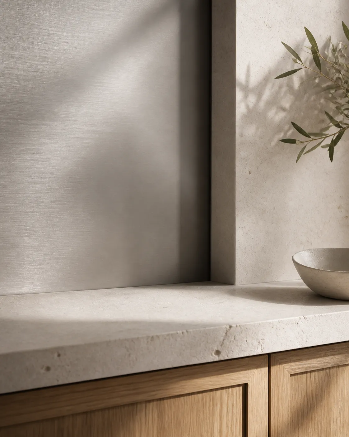 Close material study of a brushed silver-gray wall panel meeting pale limestone and warm oak cabinet fronts.